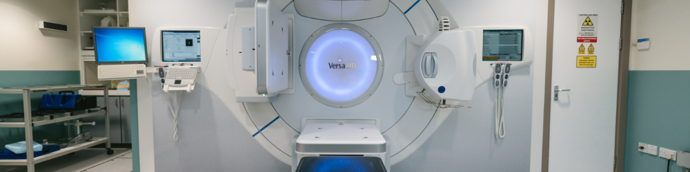 Equipamento médico Versa HD de radioterapia em sala hospitalar, com monitores e sinais de área controlada por radiação.