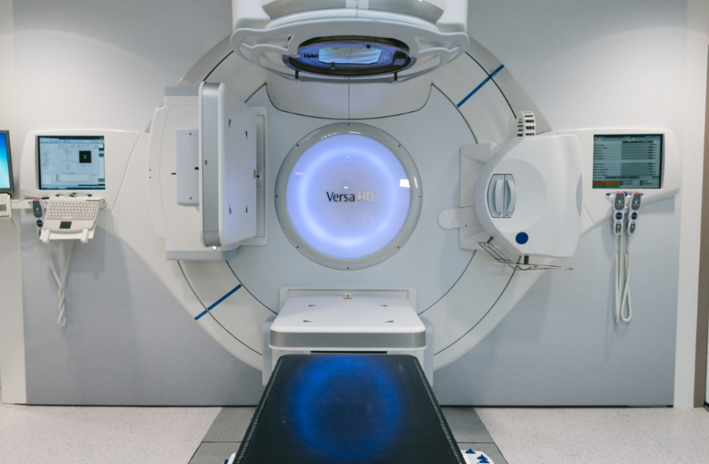 Equipamento médico Versa HD de radioterapia em sala hospitalar, com monitores e sinais de área controlada por radiação.